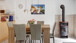 A dining area with a wooden table, grey chairs, a clock, a painting of a tropical scene, and a wood-burning stove.