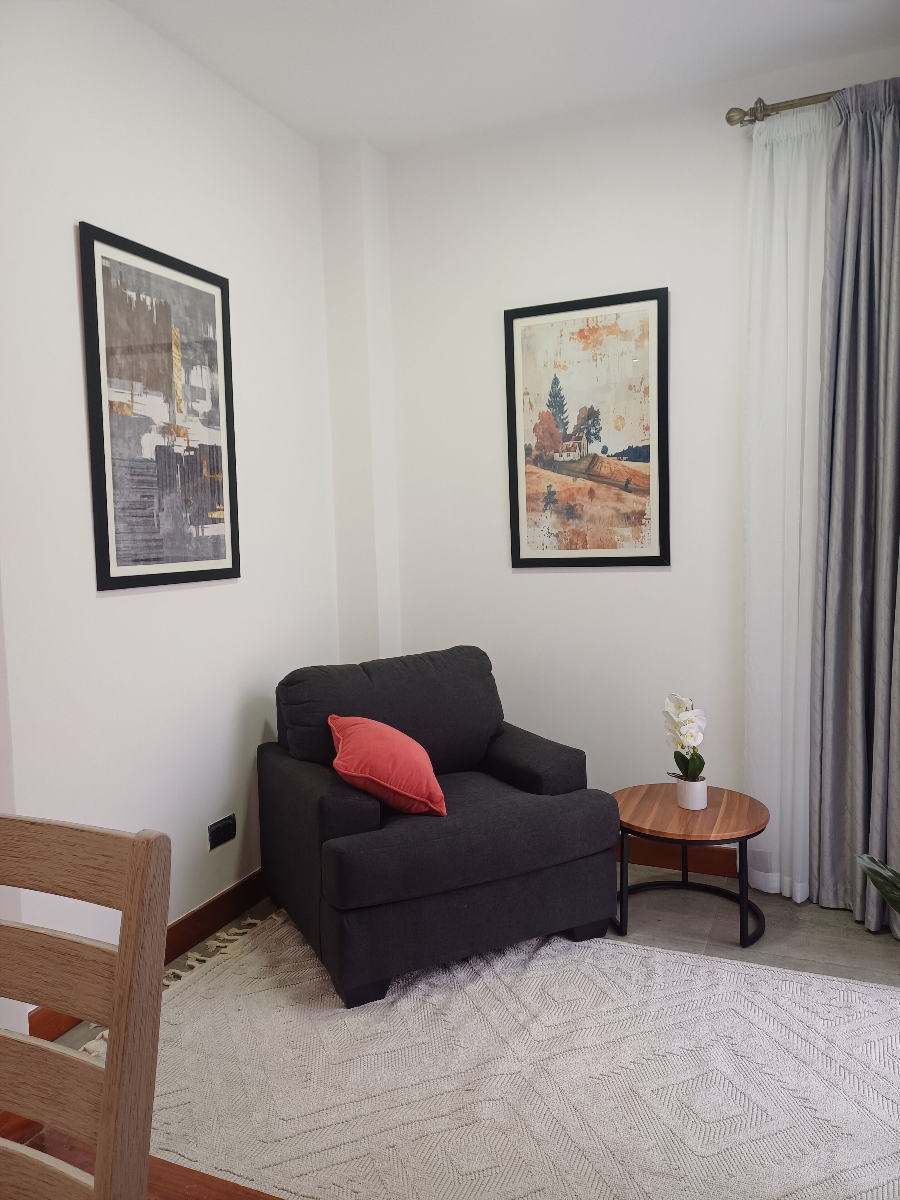 Ein Wohnzimmer mit einem schwarzen Sofa, einem roten Kissen, einem kleinen runden Holztisch mit einer weißen Blume und zwei gerahmten Bildern an der Wand.
