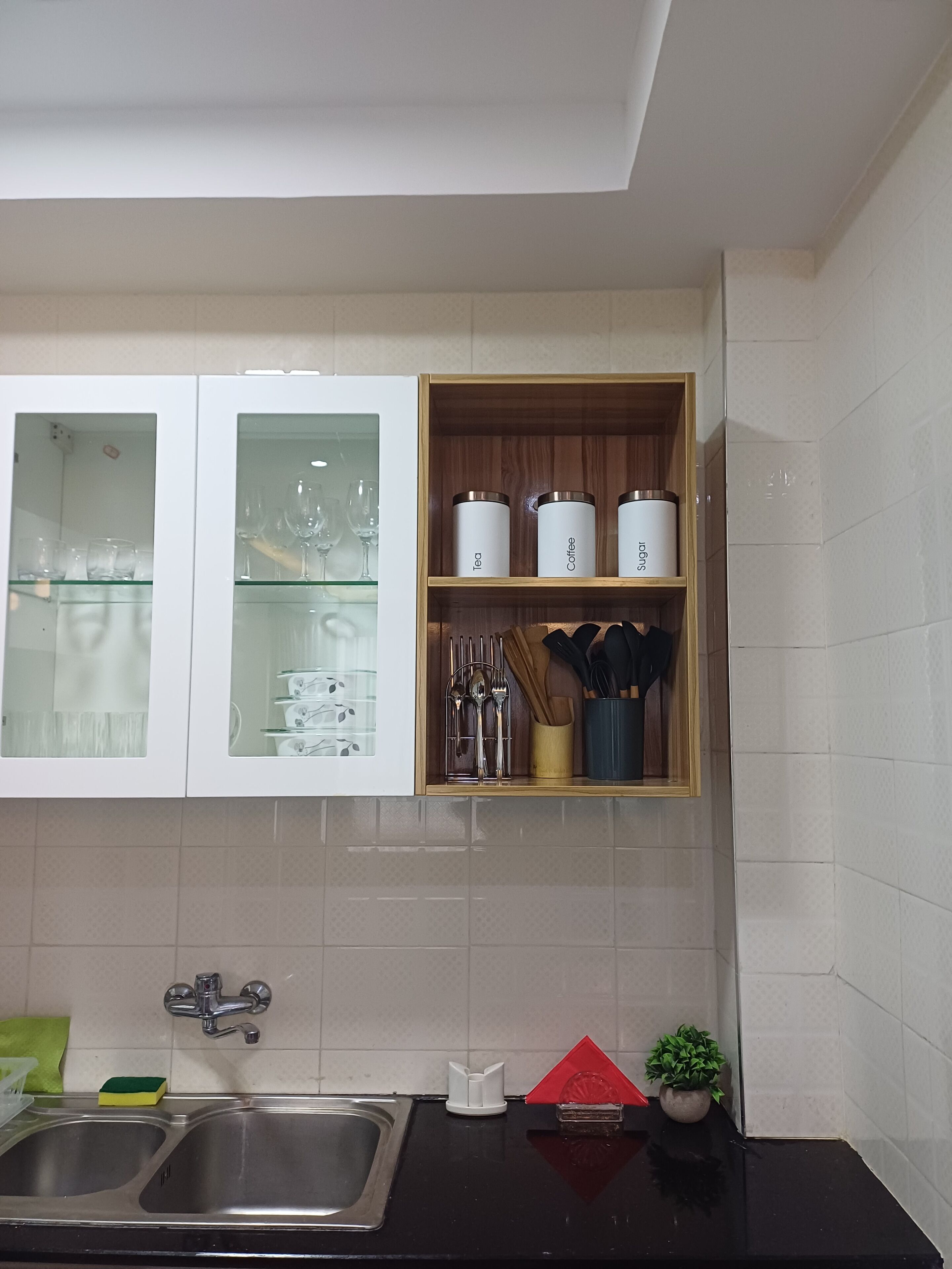 A kitchen with a double sink, a wooden shelf with kitchen utensils, and a cabinet with glass doors.