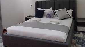 Un dormitorio moderno con una cama tapizada en gris, ropa de cama blanca, una obra de arte decorativa en la pared y un piso de parquet de madera.