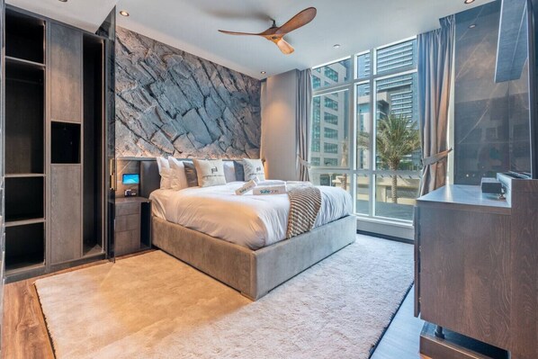 A modern bedroom with a large bed, built-in shelving, a flat-screen TV, and a city view through large windows.