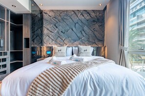 Habitación de hotel moderna con una cama grande, pared con textura de piedra y vista a un edificio.