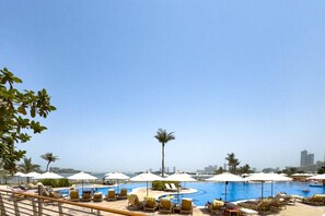 A pool area with sun loungers and umbrellas, a clear sky, and a city skyline in the background.