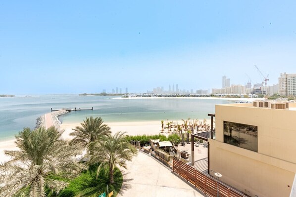 Vista de una playa con un paseo bordeado de palmeras, una playa de arena y un horizonte urbano al fondo.