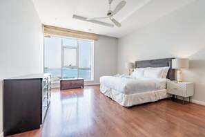 A bedroom with a large window, a bed, a nightstand, a desk, and a ceiling fan.