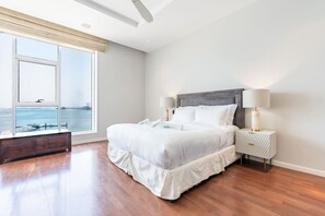 A large bed with a view of the sea through a window, a bedside table with a lamp, and a bench with a view.