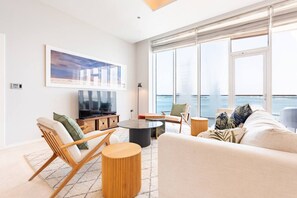 A modern living room with a large window overlooking the sea, a flat-screen TV, and a cozy seating area with a sofa and chairs.
