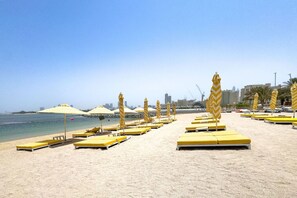 Ein Strand mit gelben Liegestühlen und gestreiften Sonnenschirmen, einem klaren Himmel und einer Stadtansicht im Hintergrund.