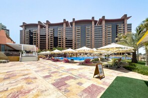 A hotel pool area with sun loungers and umbrellas, a large pool, and a building with multiple balconies in the background.