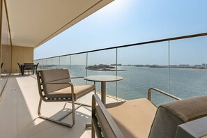 Un balcon doté d’une balustrade en verre, de deux chaises, d’une petite table ronde et offrant une vue sur la mer et le paysage urbain lointain.