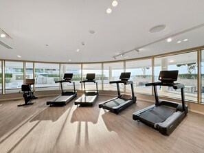 A gym with treadmills and exercise bikes, large windows, and a wooden floor.