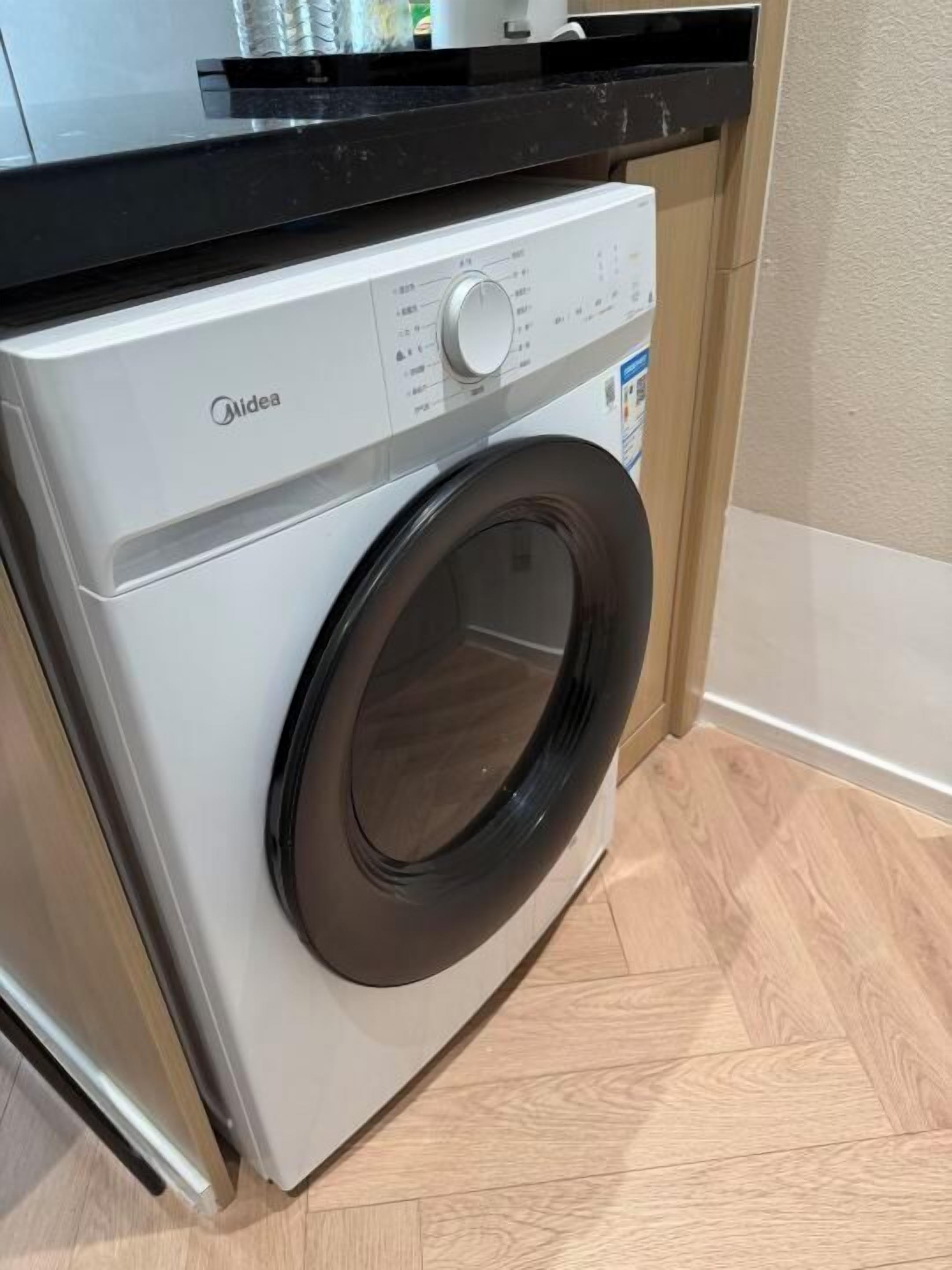 Vista frontal de una lavadora Midea en una cocina con encimera negra y suelo de madera.