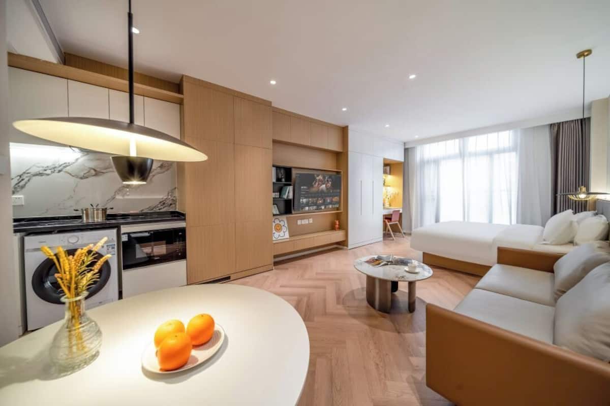 Habitación de hotel moderna con cocina, comedor y sala de estar. Incluye un sofá, una mesa de comedor con naranjas y un televisor.