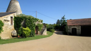 Un camino rural que lleva a un edificio de piedra con una torre cónica, rodeado de vegetación y un cielo azul despejado.