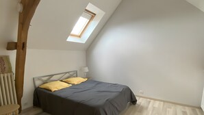 Un dormitorio con techo inclinado, tragaluz, cama con ropa oscura, mesita de noche con lámpara y radiador.