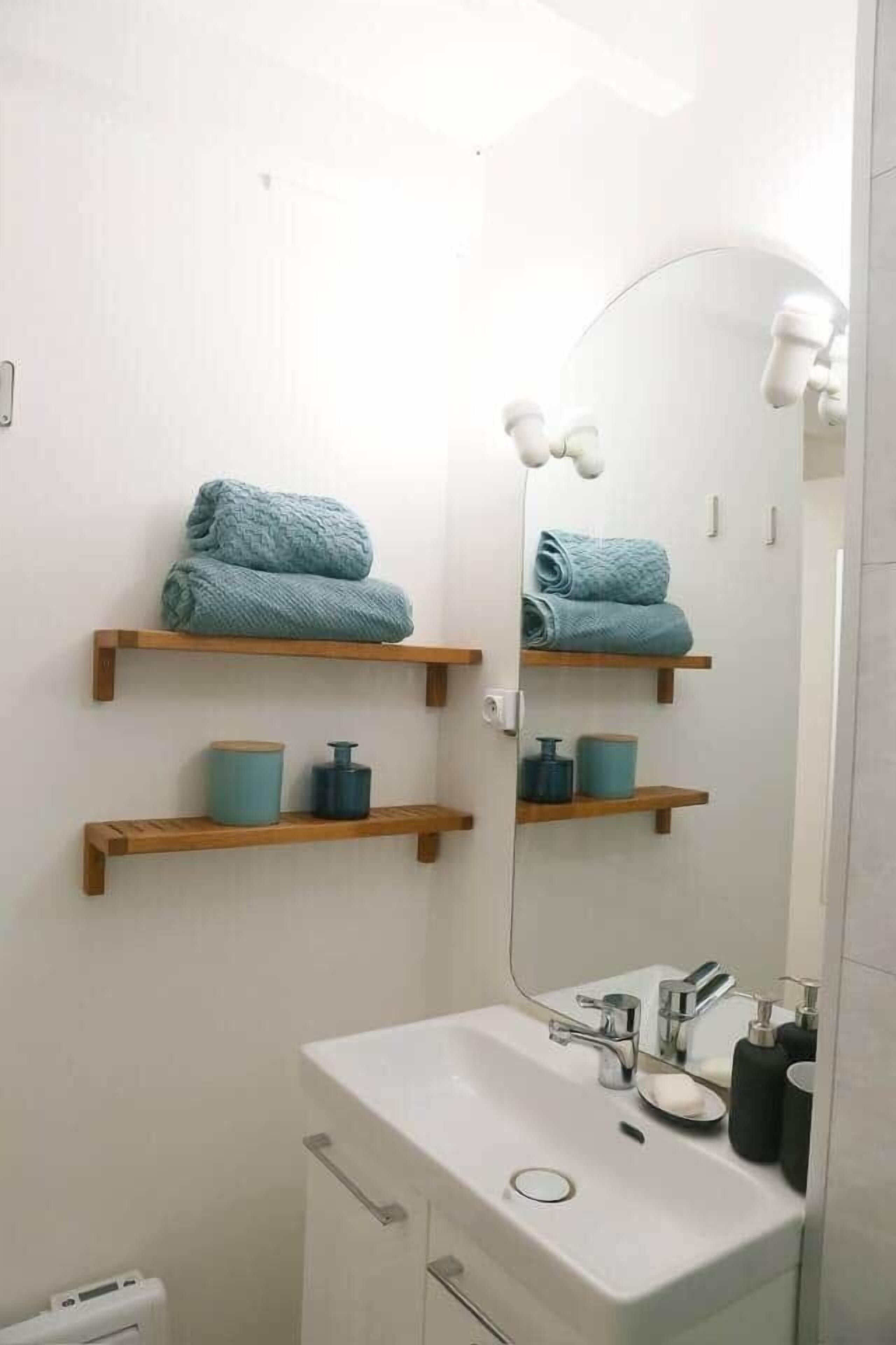 Une salle de bain avec un lavabo blanc, deux étagères en bois sur lesquelles sont posées des serviettes, et un miroir.