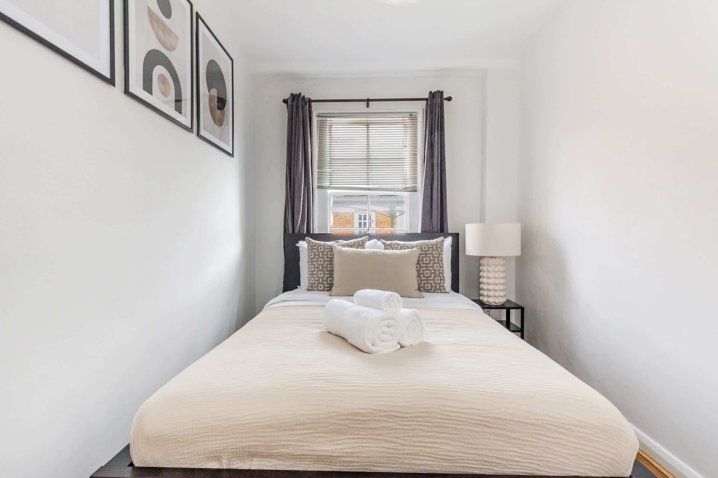 A neatly made bed with a beige bedspread and two rolled towels, a bedside table with a lamp, and a window with curtains.
