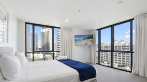 A modern hotel room with a large bed, a flat-screen TV, and expansive windows offering city views.
