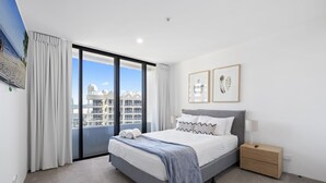 A modern hotel room with a large bed, a nightstand, and a view of the cityscape through floor-to-ceiling windows.