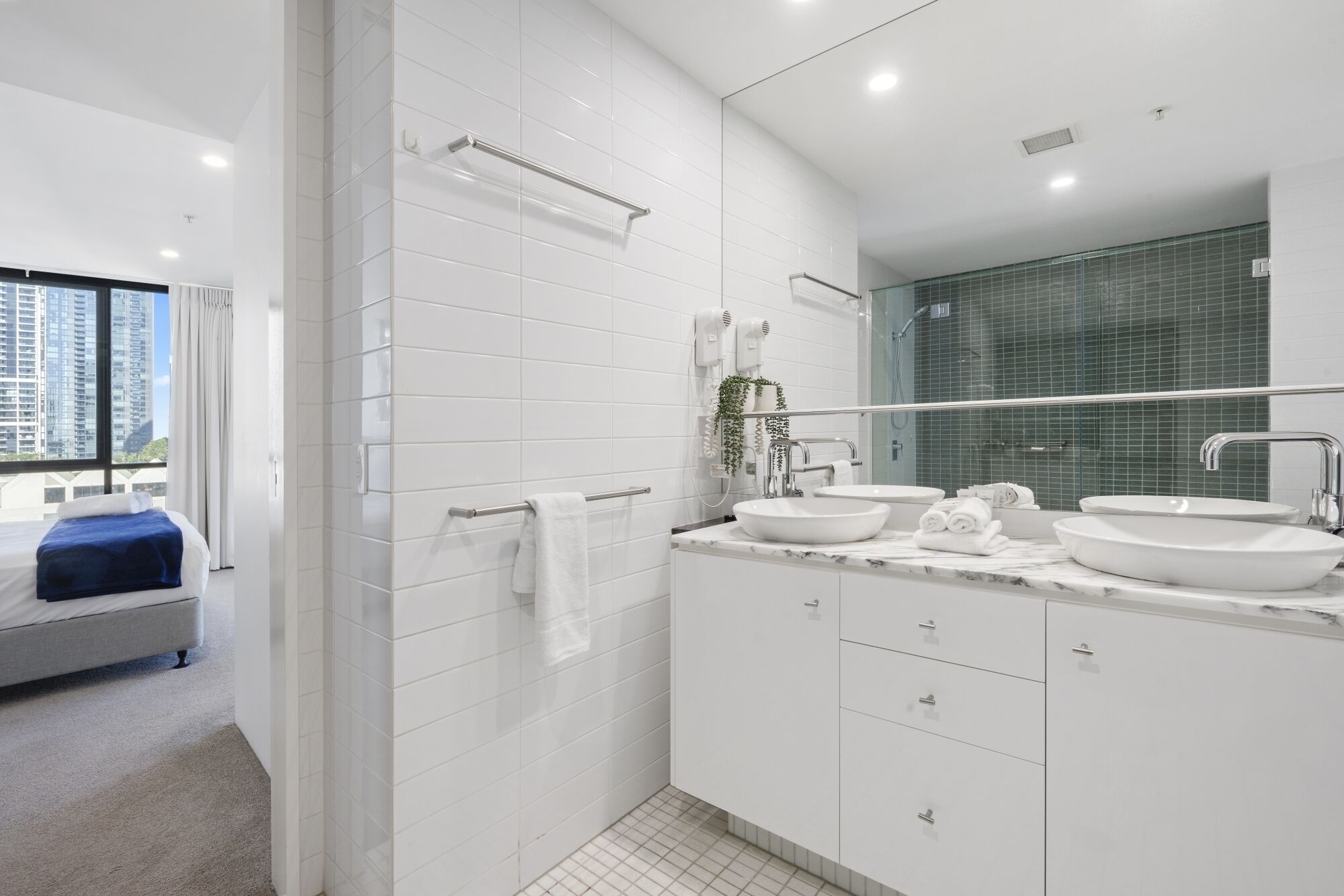 A modern bathroom with white tiles, a large mirror, two sinks, and a shower.