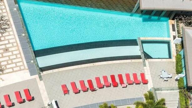 A rooftop pool with a clear blue water feature, surrounded by red lounge chairs and a small seating area.