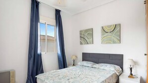 A bedroom with a bed, two wall art pieces, a nightstand, a lamp, and a window with curtains. - Beautiful 3-bedroom home in Playa Flamenca, Alicante. Spain. (Orihuela)