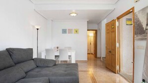 A living room with a grey sofa, a white coffee table, and a dining area with a white table and chairs. There is a hallway leading to a door with a poster on the wall. - Beautiful 3-bedroom home in Playa Flamenca, Alicante. Spain. (Orihuela)
