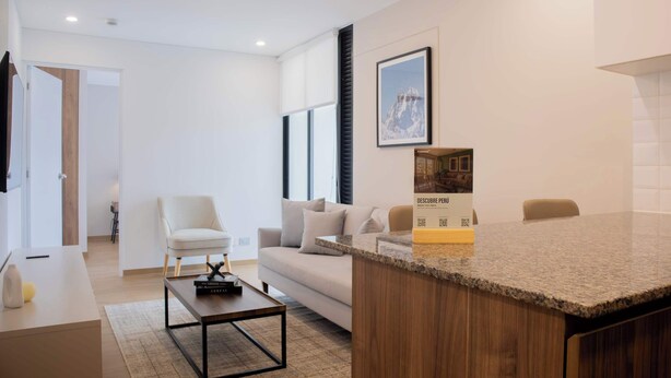A modern living room with a sofa, a coffee table, and a white armchair. There is a framed picture on the wall and a small table with a book and a bottle. - w* | Chic 1BR Apartment in Miraflores (Lima)