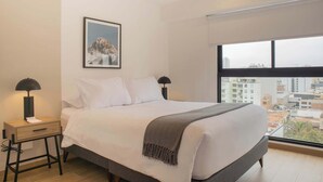 A modern bedroom with a large bed, a nightstand, a lamp, a framed picture on the wall, and a city view through the window. - w* | Chic 1BR Apartment in Miraflores (Lima)