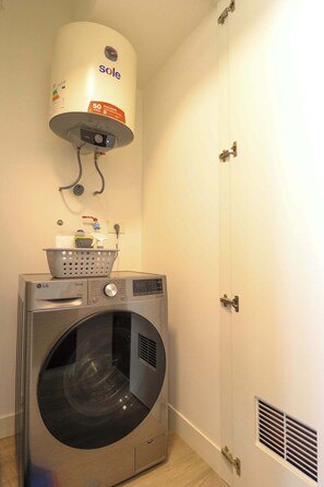 Une petite buanderie équipée d’un lave-linge à chargement frontal, d’un chauffe-eau mural et d’un panier posé sur le lave-linge.