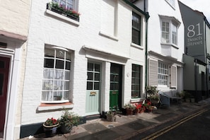 Um edifício branco com porta verde e várias janelas. Há plantas em vasos na calçada e uma placa indicando "81 Beach Street".
