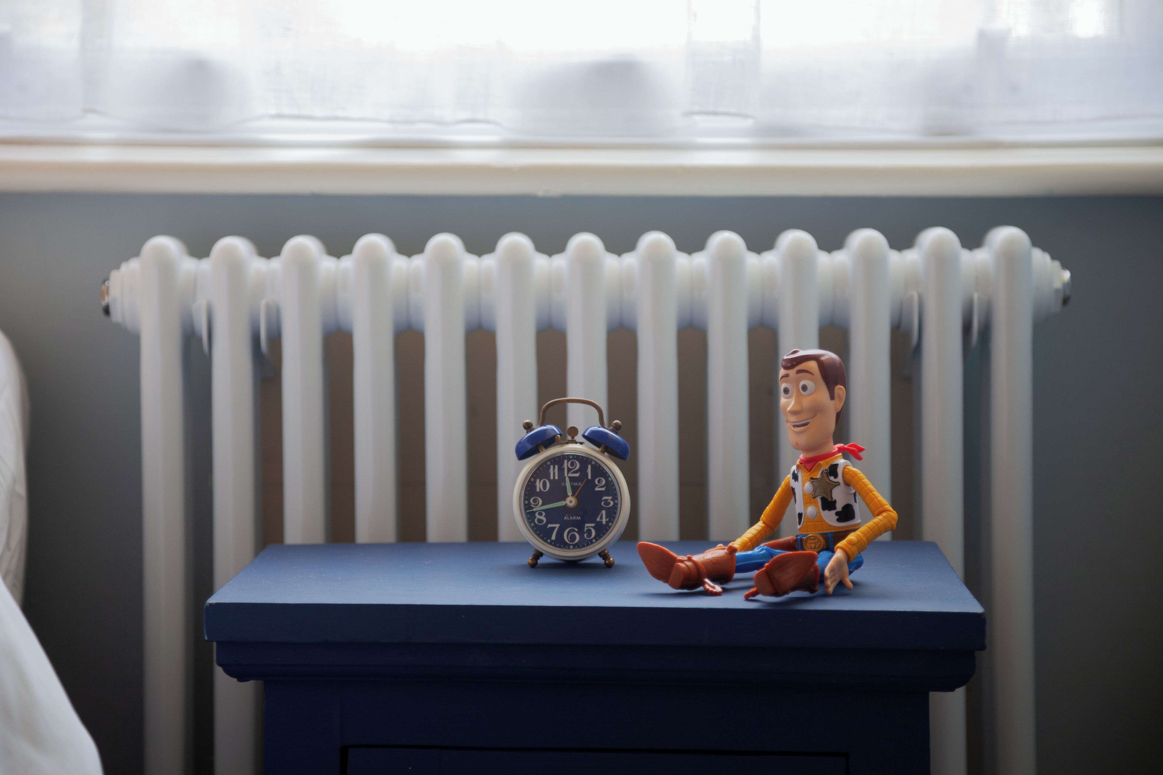 A toy figure sitting on a blue table with a clock beside it, in front of a radiator.