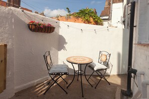 Um pequeno pátio com uma mesa redonda e duas cadeiras, cercado por paredes brancas e uma cesta de flores suspensa.