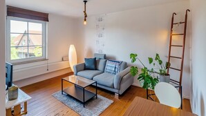 Ein modernes Wohnzimmer mit einem grauen Sofa, einem Glastisch, einer weißen Lampe, einer Holzleiter und einem Blumentopf.