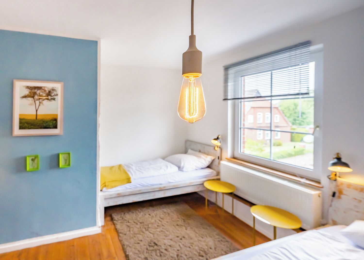 Une chambre à coucher avec un lit, un luminaire suspendu, une fenêtre avec des stores et un tableau encadré au mur.