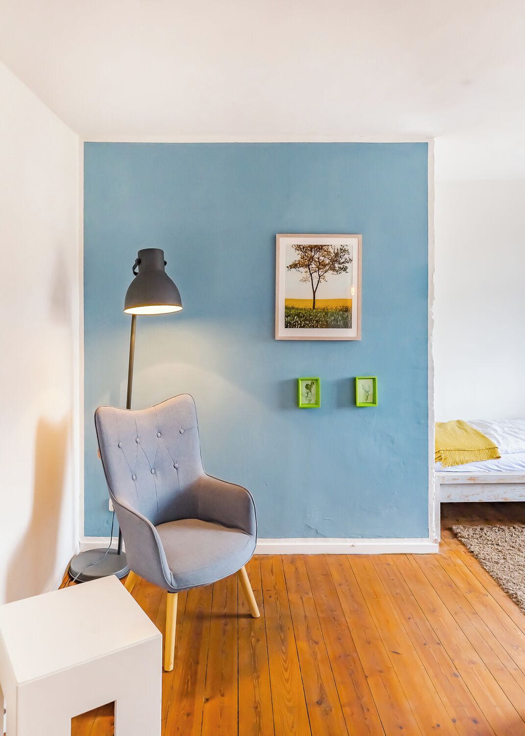 Una habitación con pared azul, una butaca tapizada en gris, una lámpara de pie, una cama con manta amarilla y un cuadro con una imagen de un árbol.