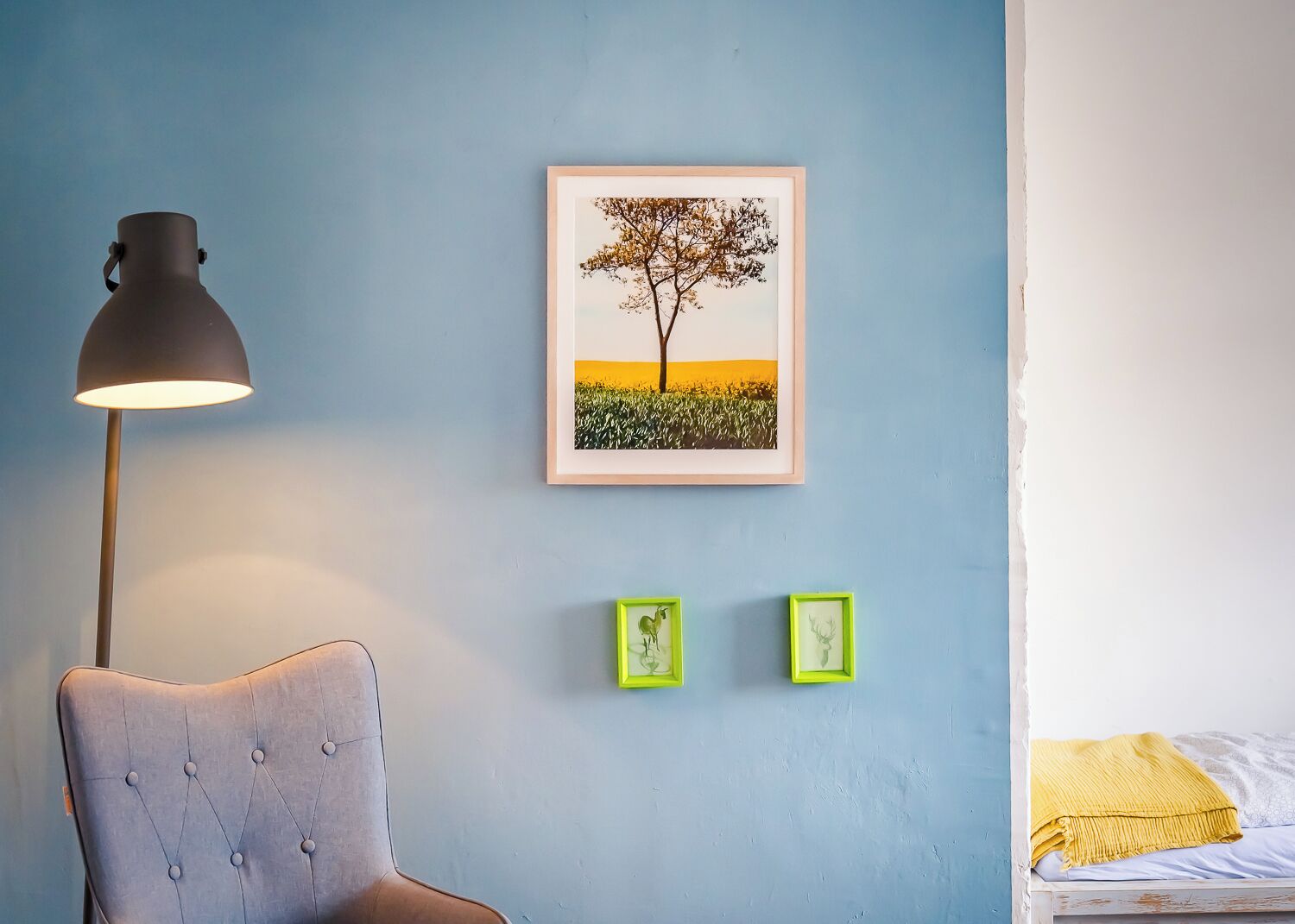 Ein Zimmer mit blauer Wand, einem gerahmten Bild von einem Baum, einer Lampe und zwei kleinen Bildern in grünem Rahmen.