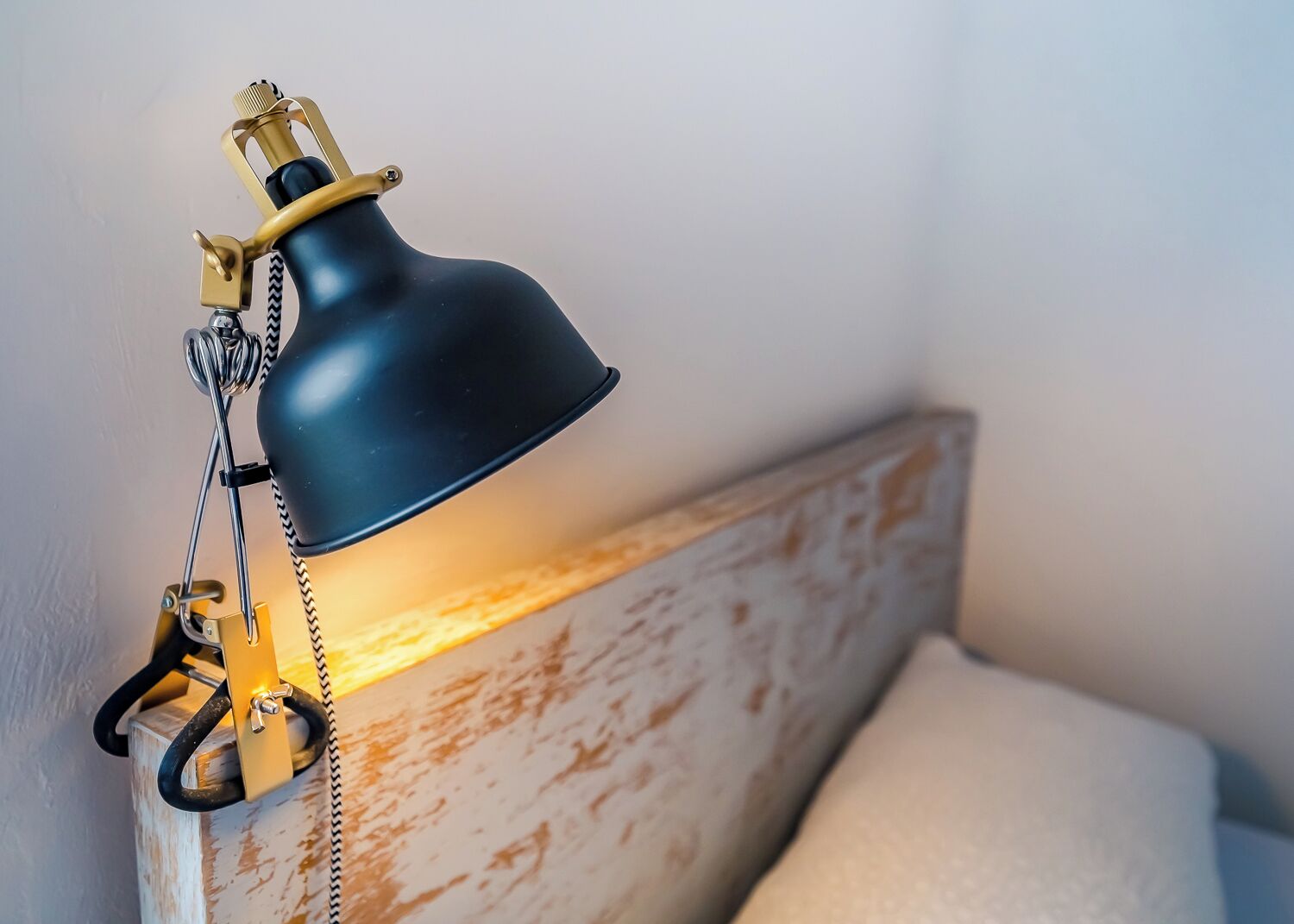 Uma luminária de mesa com cúpula preta e detalhes dourados, fixada em uma cabeceira de madeira com acabamento desgastado.