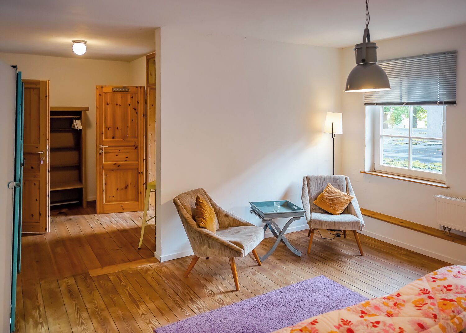 Una habitación con piso de madera, dos sillones, una mesita, una cama con ropa de cama floral y una ventana con persianas, además de una lámpara en el techo.