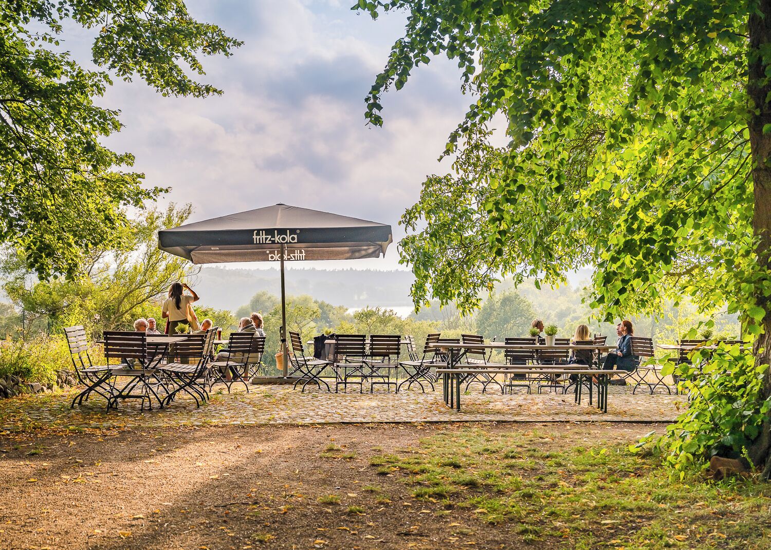 An outdoor seating area with tables and chairs under a large umbrella, surrounded by trees and a view of the landscape.