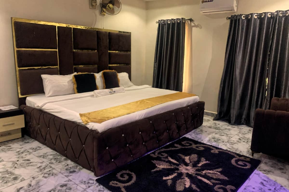 Un dormitorio con una cama grande, cabecera con detalles dorados, una mesita de noche, un ventilador fijado a la pared y una ventana con cortinas.