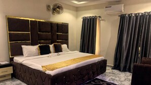A bedroom with a large bed, a headboard with a gold trim, a bedside table, a wall-mounted fan, and a window with curtains.