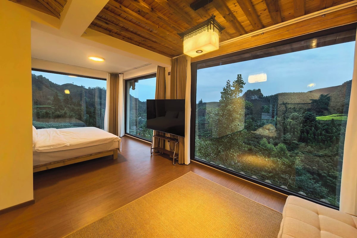 A modern hotel room with a large bed, a TV, and a view of a forested landscape.