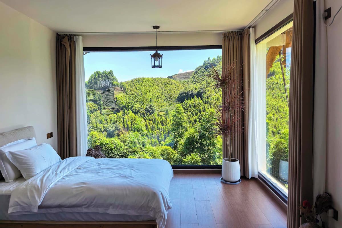 A bedroom with a large window offering a view of a lush green forest and hills.