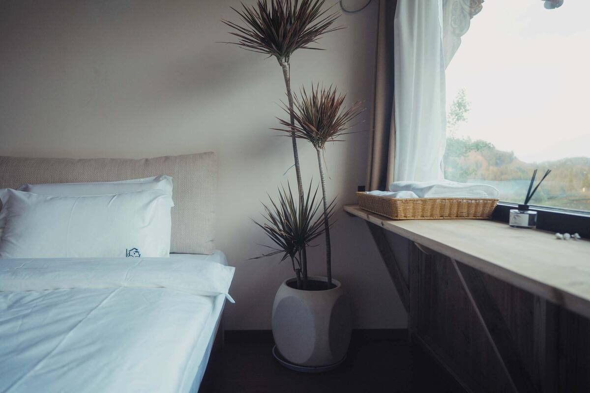 A neatly made bed with white linens, a tall potted plant beside it, and a window with a view of a landscape.