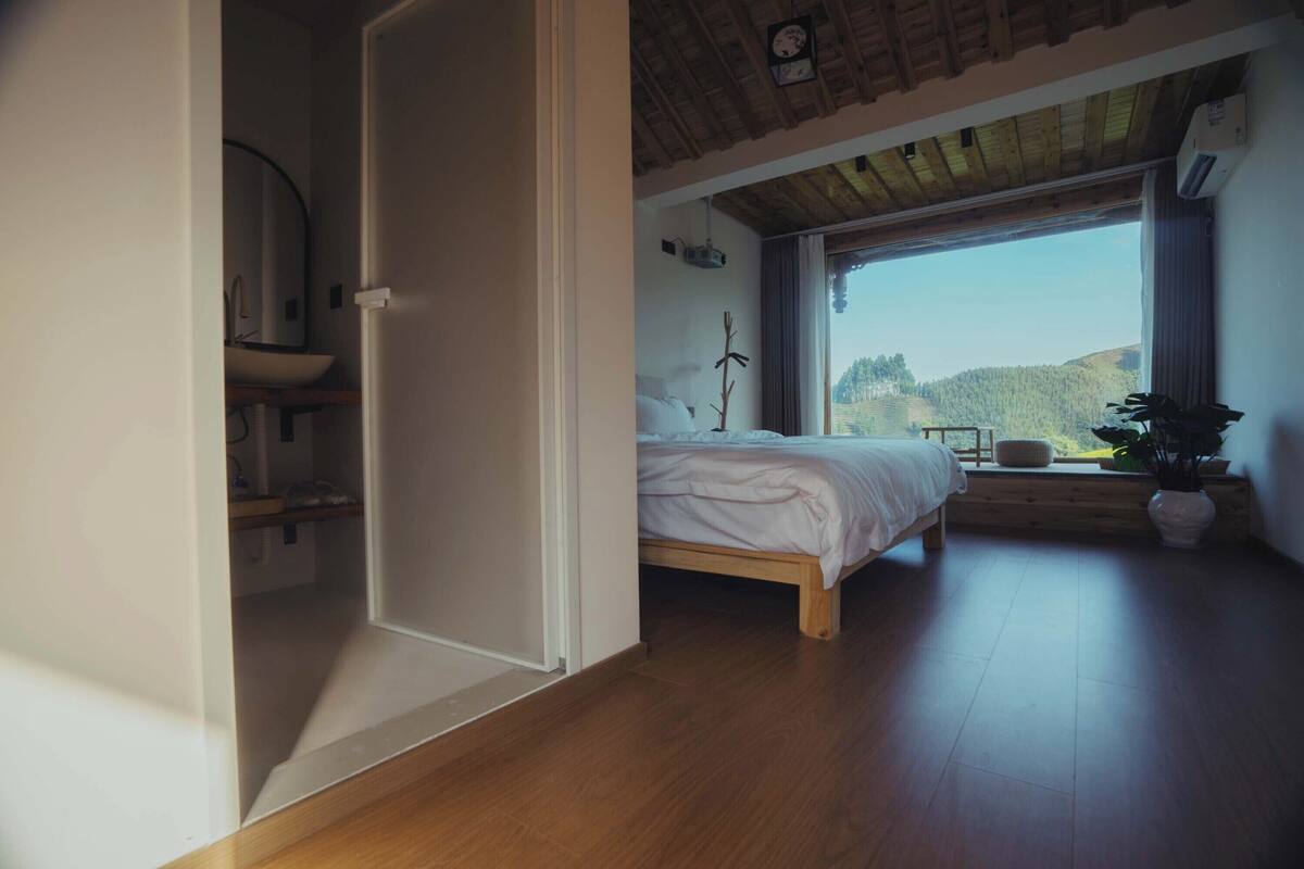 A bedroom with a wooden bed, a window with a view of hills, and a bathroom with a sink and mirror.