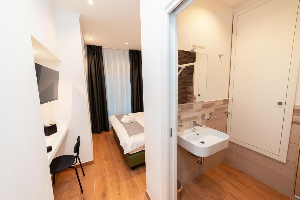 A modern hotel room with a bed, a small desk, a chair, and a bathroom with a sink and mirror.