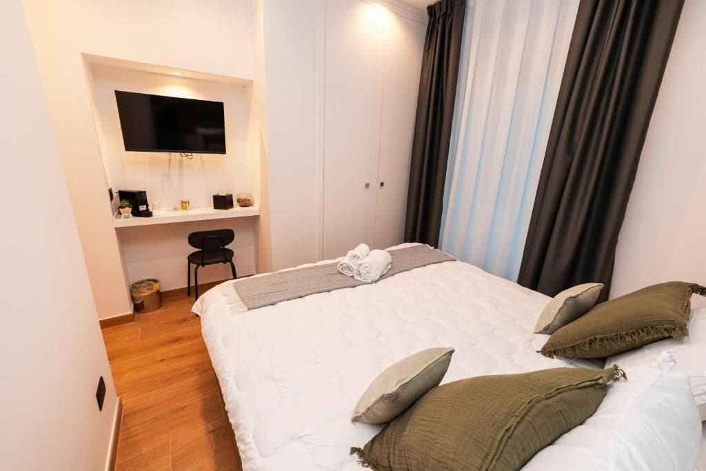 A bedroom with a bed, a TV mounted on the wall, a desk with a chair, and a window with curtains.