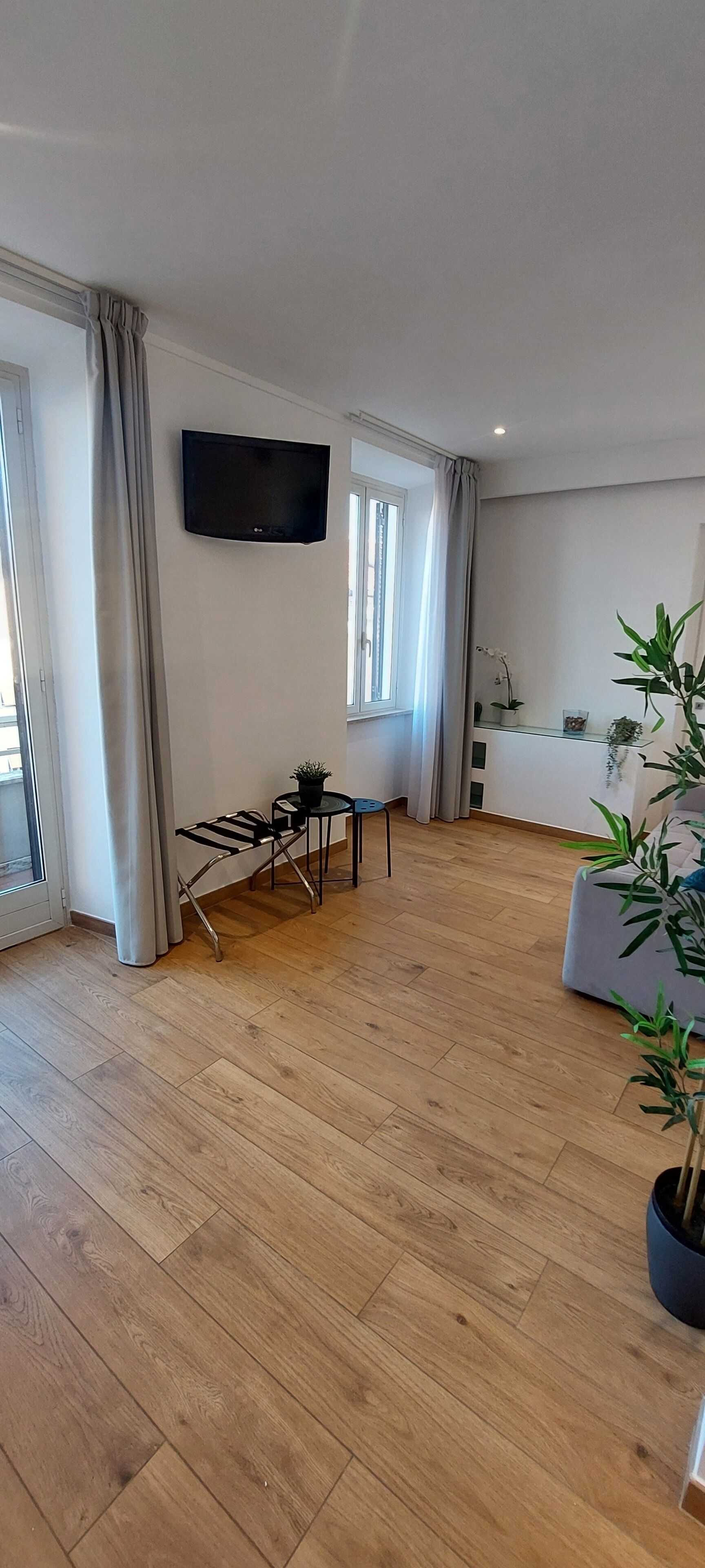 A spacious living area with a flat-screen TV mounted on the wall, a dining table with chairs, and a potted plant.