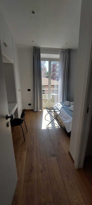 A bedroom with a bed, a chair, a sliding glass door leading to a balcony, and a wooden floor.